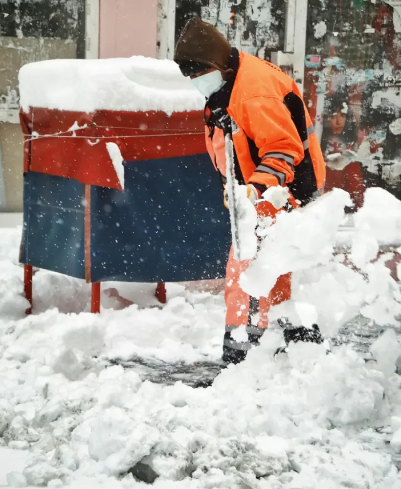 A person in orange winter workwear and a mask shovels snow on a city sidewalk during heavy snowfall, performing essential exterior cleaning.